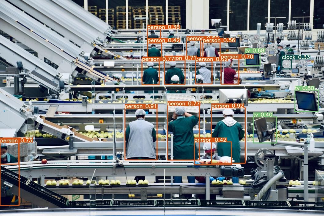 Linea di produzione food vista dall'alto con sistema AI che identifica simultaneamente decine di operatori alle postazioni di lavoro tramite bounding box arancioni con label 'person' e percentuale di confidenza